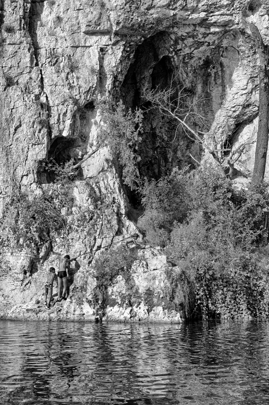 Trois silhouettes minuscules remontent une paroi rocheuse le long d'un câble, en diagonale du cadre.