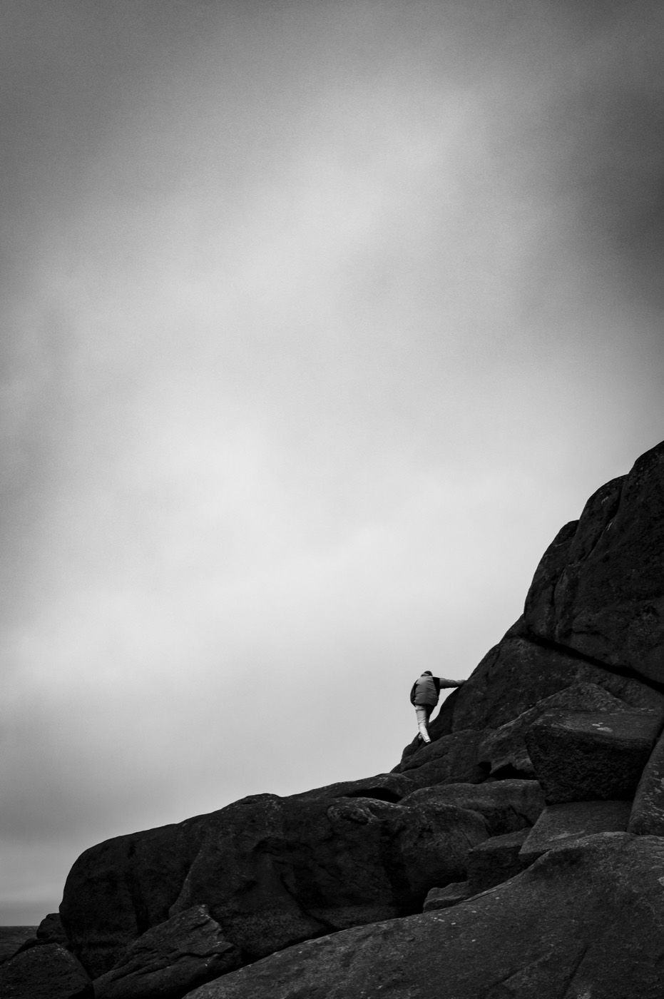 Une silhouette gravit un chaos de blocs de granite, seule contre un ciel entièrement vide. La roche occupe le bas du cadre, tout le reste est du gris.