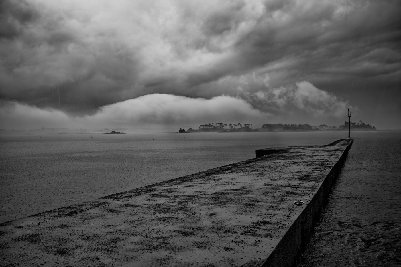 La jetée s'avance dans l'eau grise vers l'île, tandis qu'un front d'orage écrase l'horizon et qu'un rideau de pluie efface la moitié du paysage. Un mât se dresse à droite, seul point vertical du cadre.