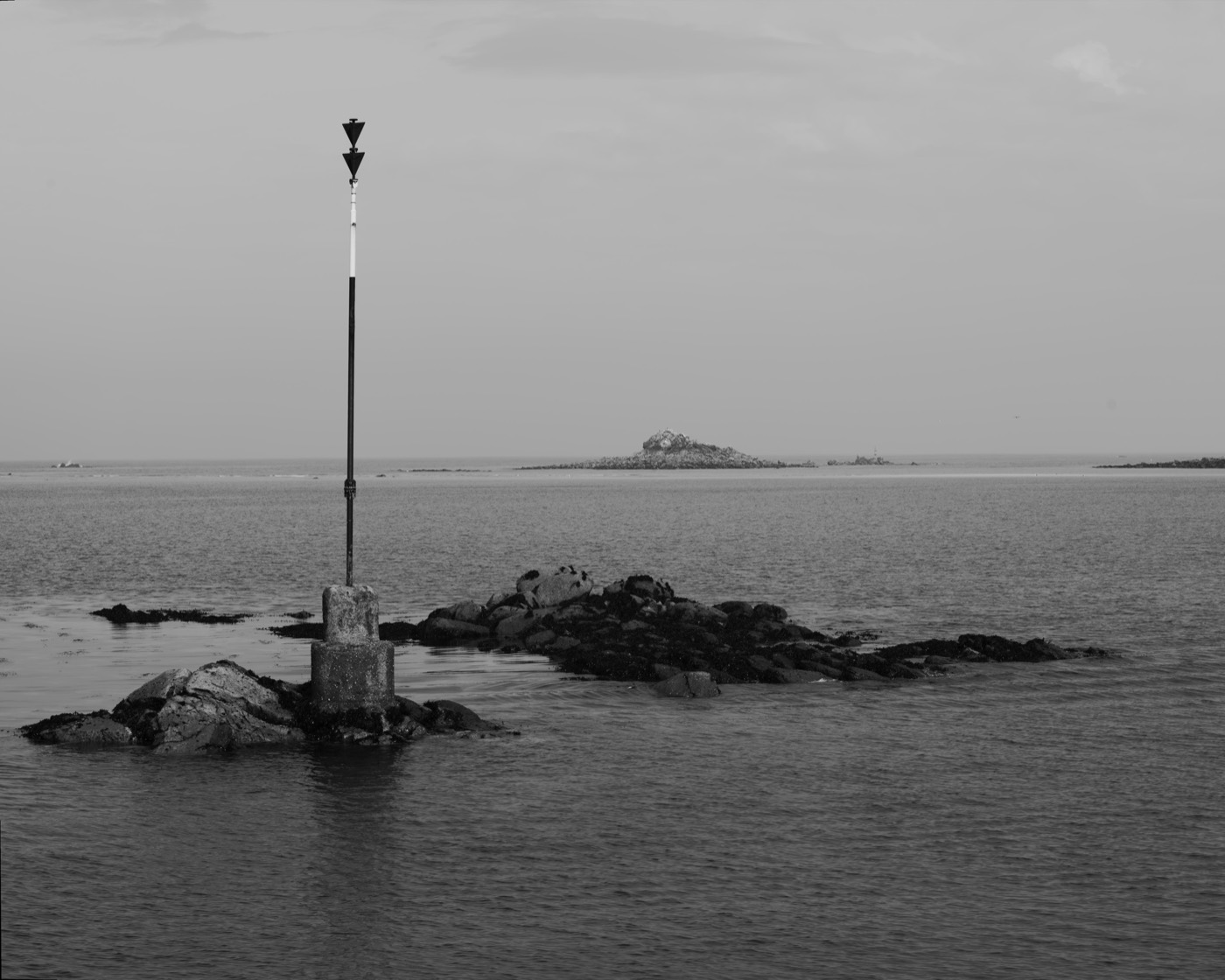 Une perche cardinale noire sur un socle de béton clair, posée sur une roche que la marée découvre ; la mer et le ciel ont la même valeur de gris.