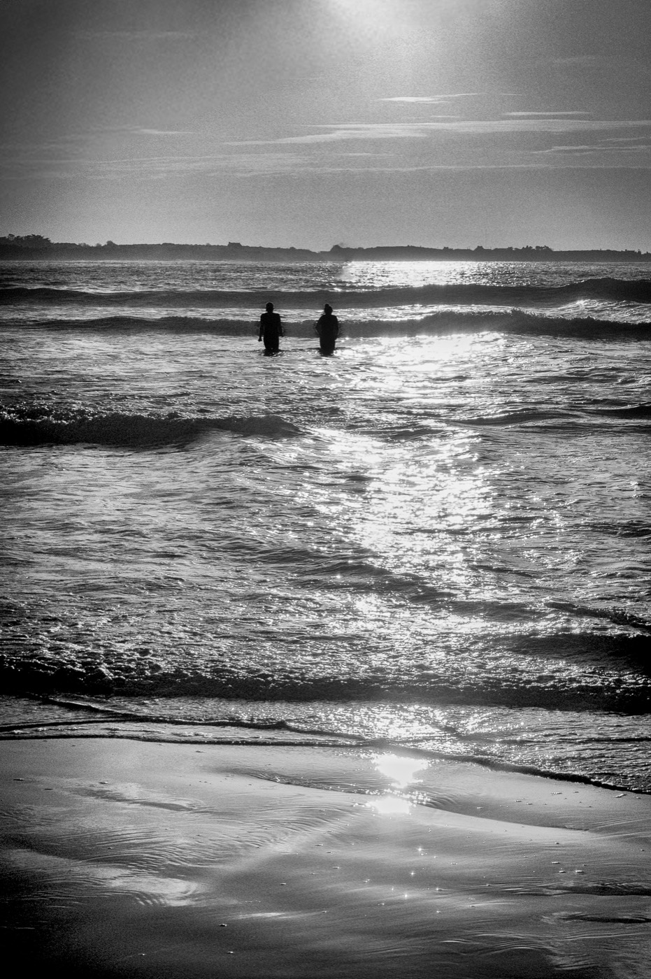 Deux silhouettes debout dans les vagues, à contre-jour, sur une traînée de lumière qui court jusqu'au rivage.