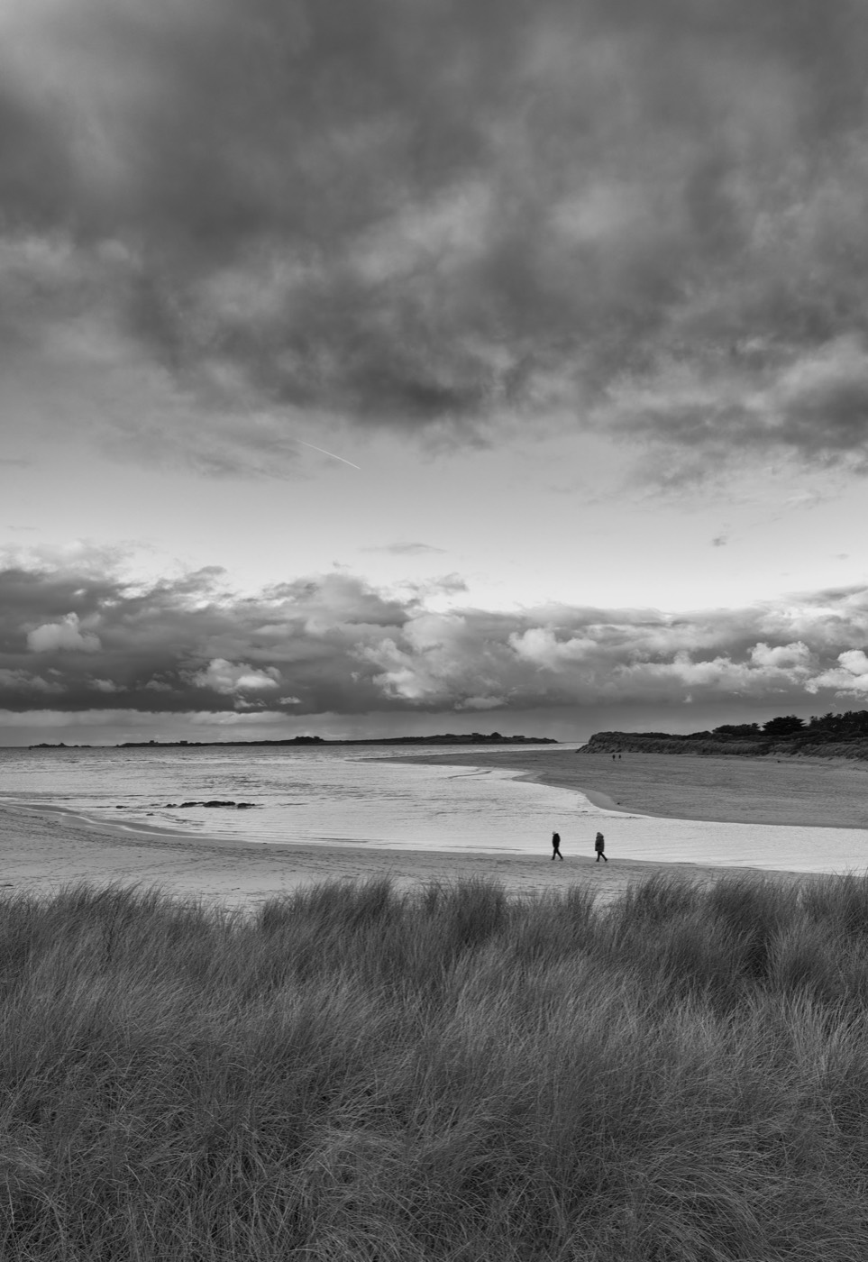 Deux marcheurs minuscules traversent une plage vide, séparés du regard par une dune d'herbes sèches. Le ciel occupe les deux tiers de l'image.