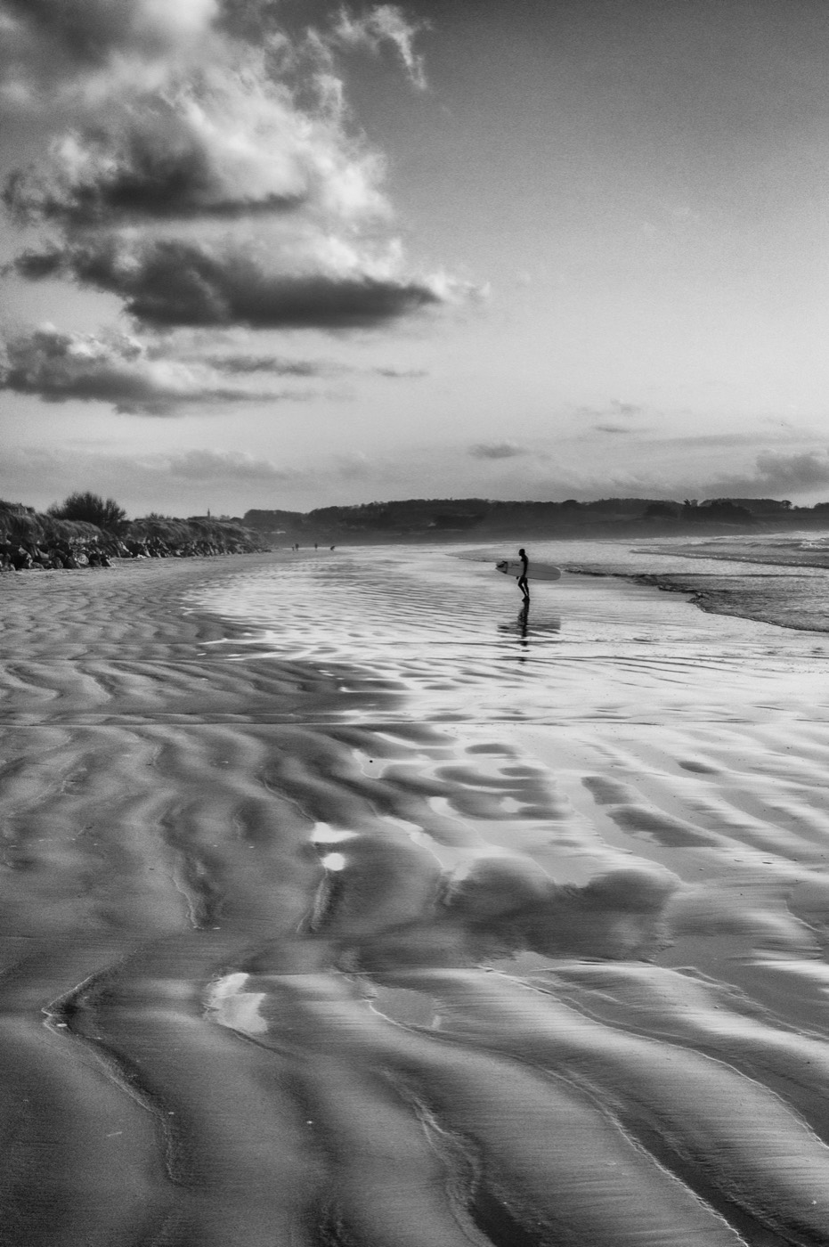Une silhouette portant une planche traverse l'estran, seule au milieu des ondulations que la marée a laissées dans le sable.