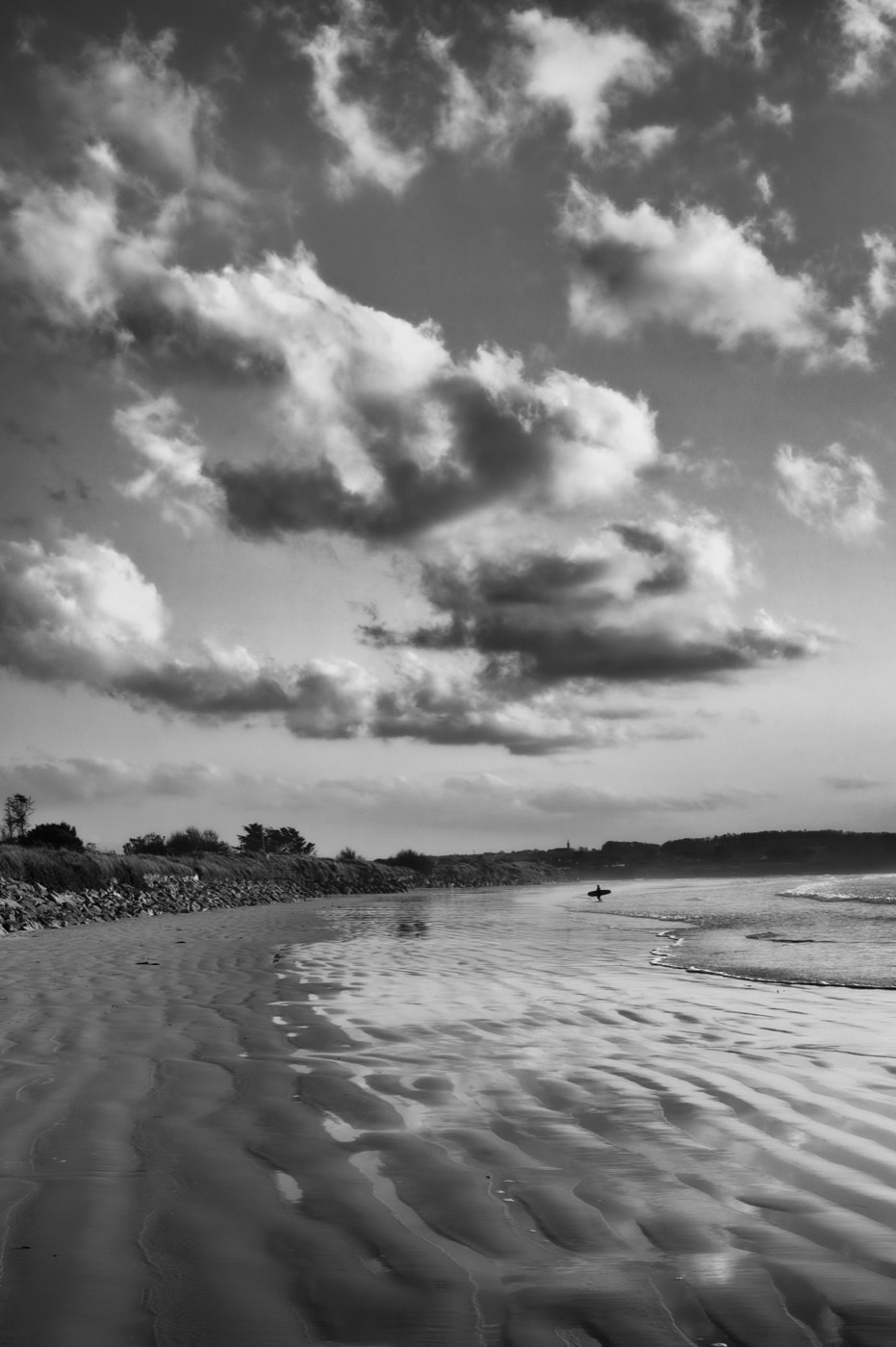 Le sable mouillé garde les ondulations laissées par la marée ; une silhouette portant une planche marche au loin. Le ciel est plein de nuages ronds.