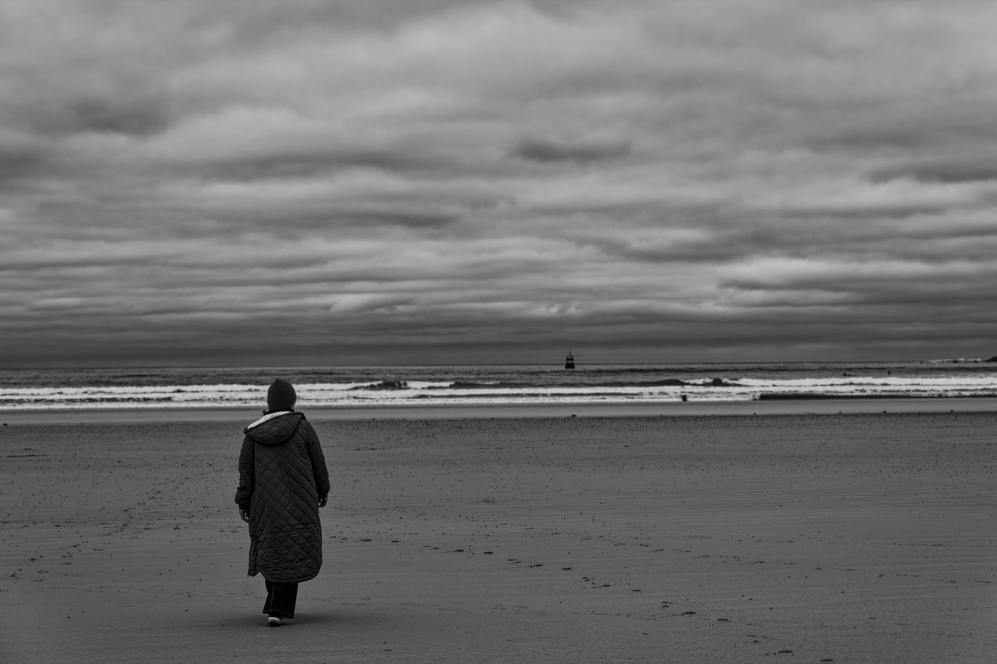 Une silhouette en manteau marche sur l'estran, l'horizon très bas, une balise minuscule au loin.