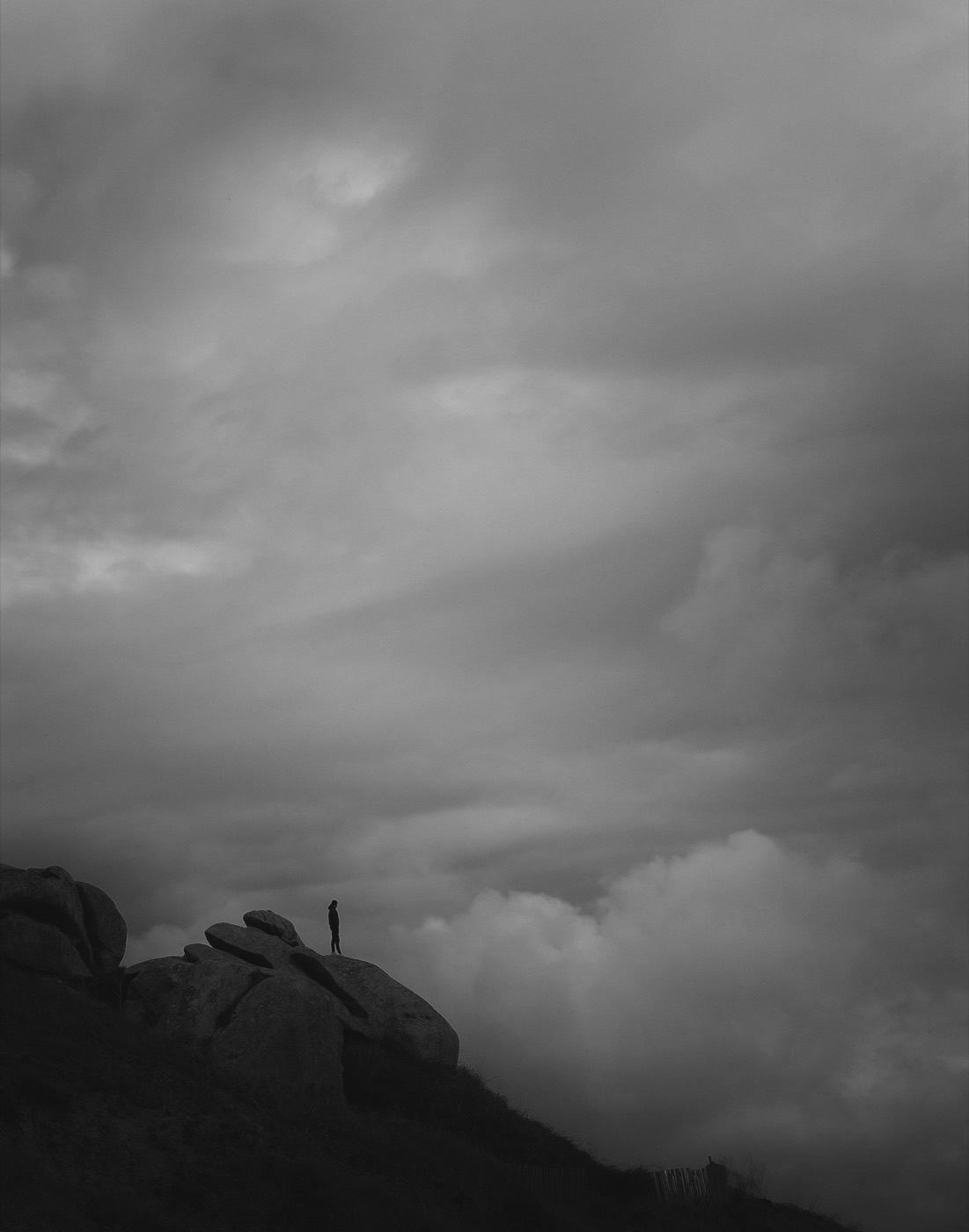 Une falaise sombre en diagonale, une silhouette minuscule à son bord, la brume au-dessous.
