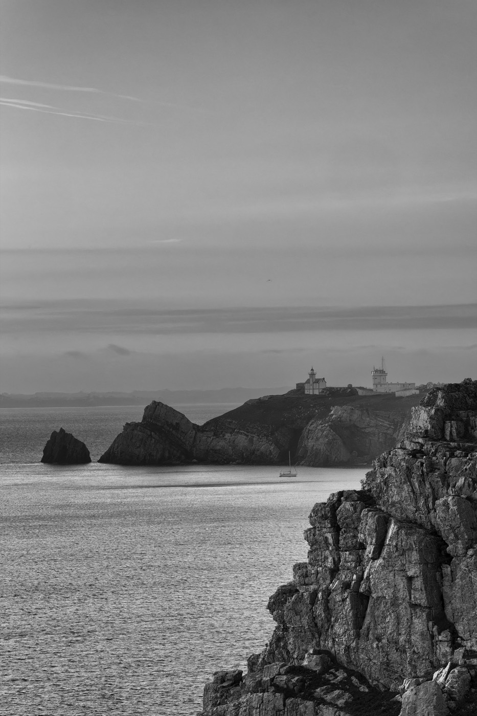 Une falaise sombre au premier plan, un phare posé sur la pointe au loin, la mer argentée entre les deux.