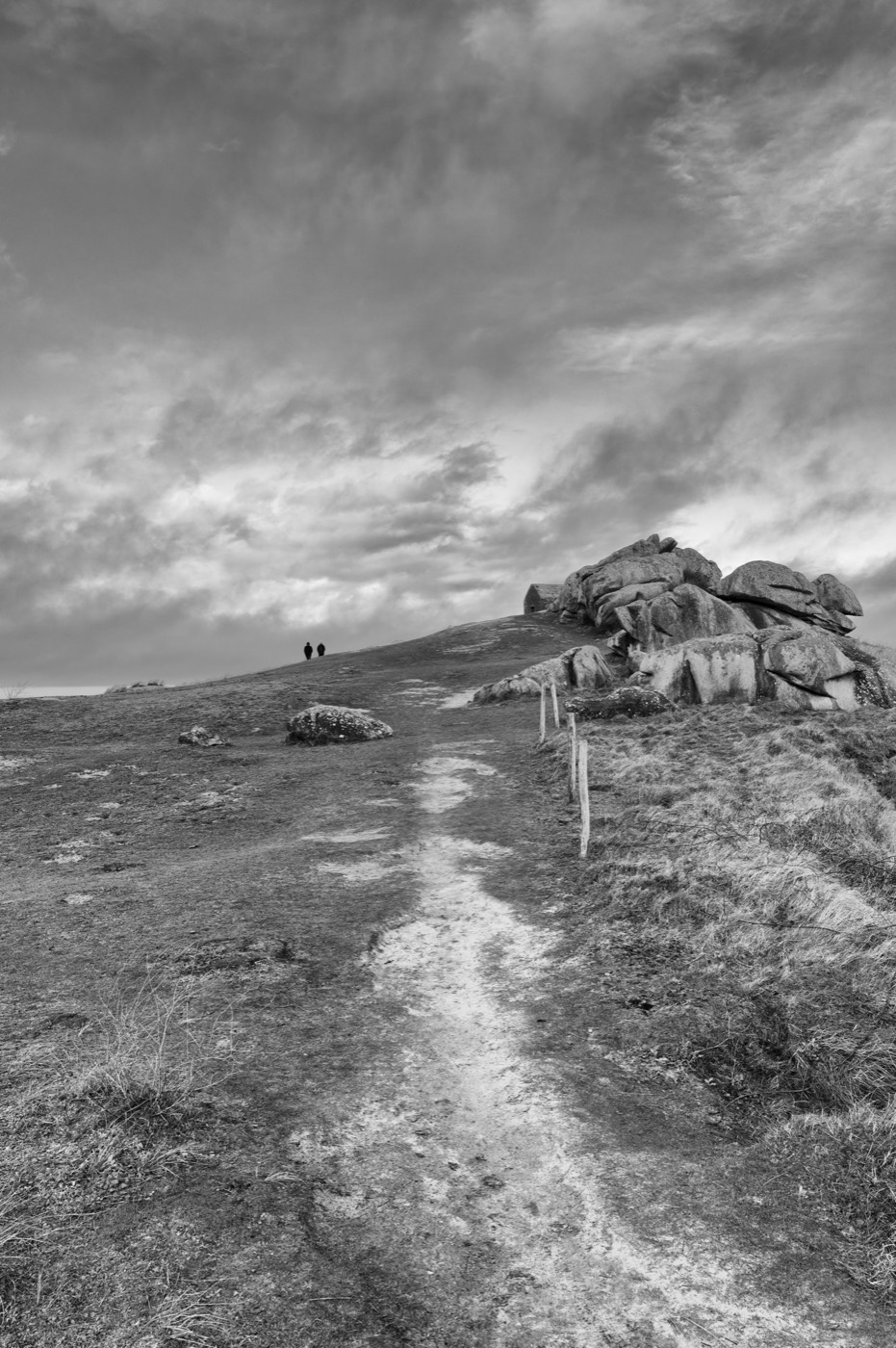 Un sentier de sable monte vers la crête, où deux silhouettes se découpent contre le ciel. À droite, un chaos de blocs de granite tient le haut de la pente.