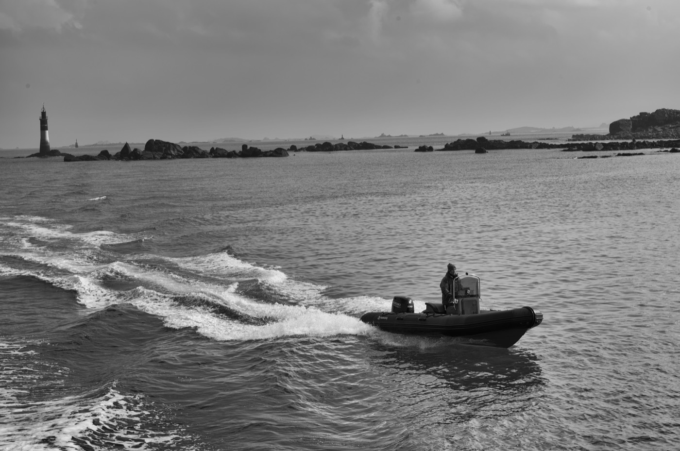 Une silhouette debout à la barre d'un bateau, capuche et veste sombre, une main sur la console.