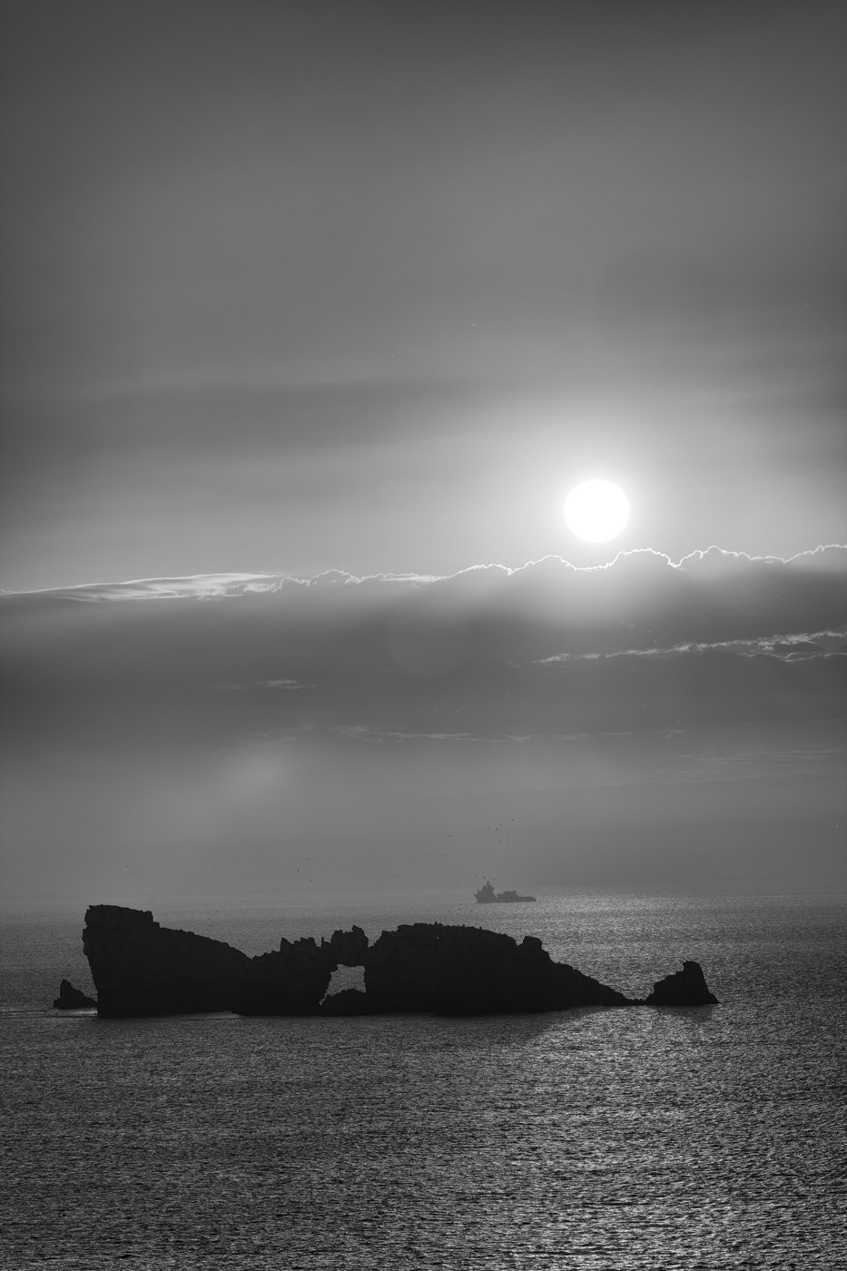 Un îlot sombre percé d'une arche, posé sur la mer, le soleil bas juste au-dessus.