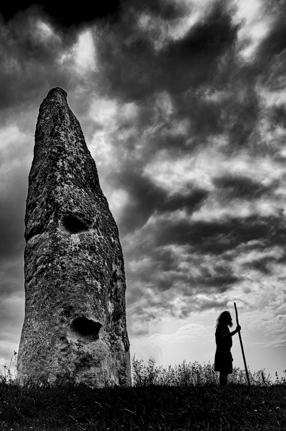 Une pierre dressée vue en contre-plongée, le ciel déchiré derrière elle ; une silhouette debout à sa droite, un bâton à la main.