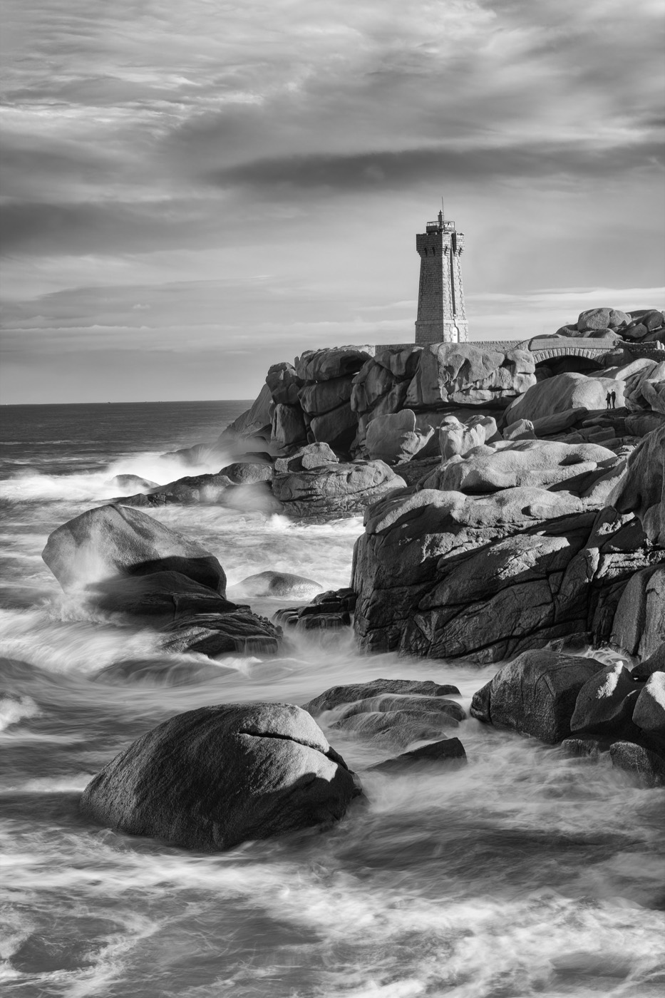 Un phare sur ses rochers, la mer lissée par une pose longue, le ciel chargé.