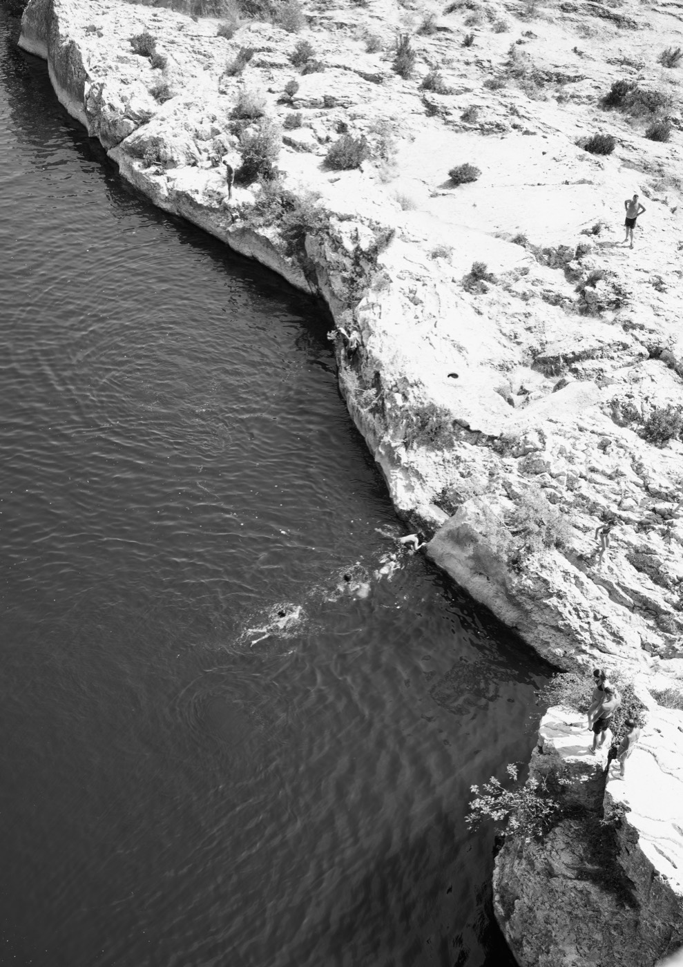 Vue de dessus : la roche occupe la moitié droite du cadre, l'eau l'autre moitié, des silhouettes réparties le long de la diagonale qui les sépare.
