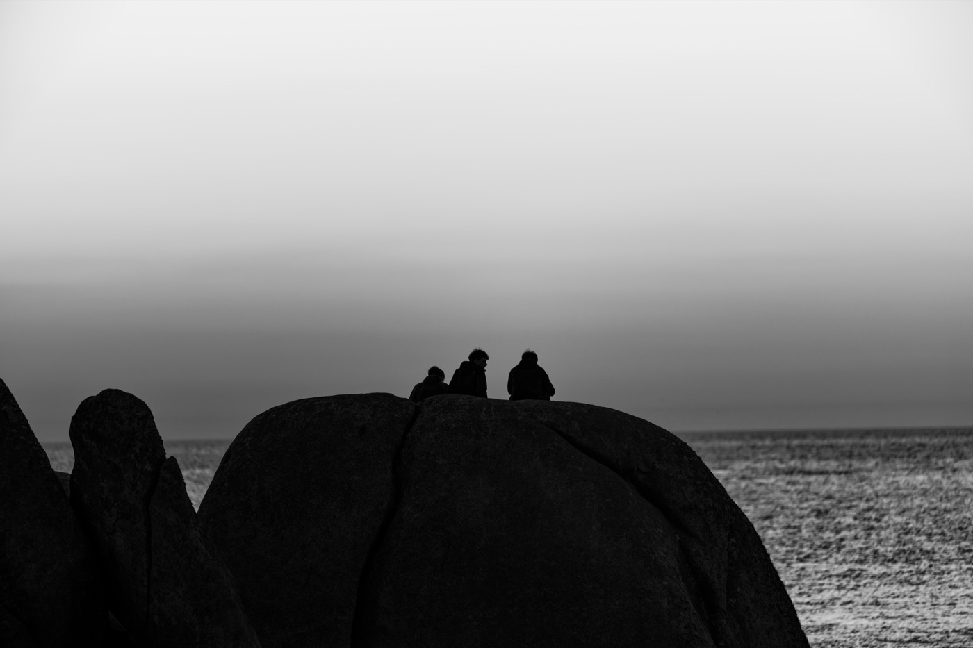 Deux silhouettes assises au sommet d'un rocher sombre, en contre-jour sur un ciel blanc.